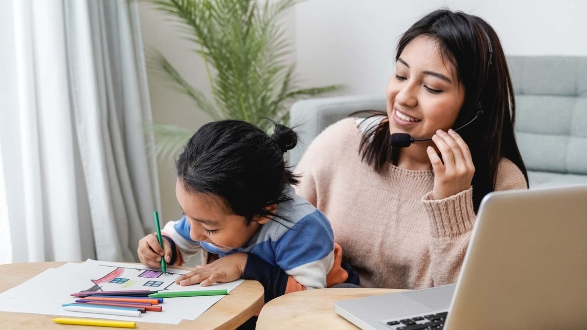 A mother working on a laptop beside her child who is drawing